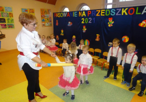 Pani dyrektor Maria Królikowska trzyma w prawej dłoni dużą żółtą kredką, którą przykłada dziewczynce do lewego ramienia. W lewej dłoni trzyma dłoń dziewczynki. W tle stoją dzieci.